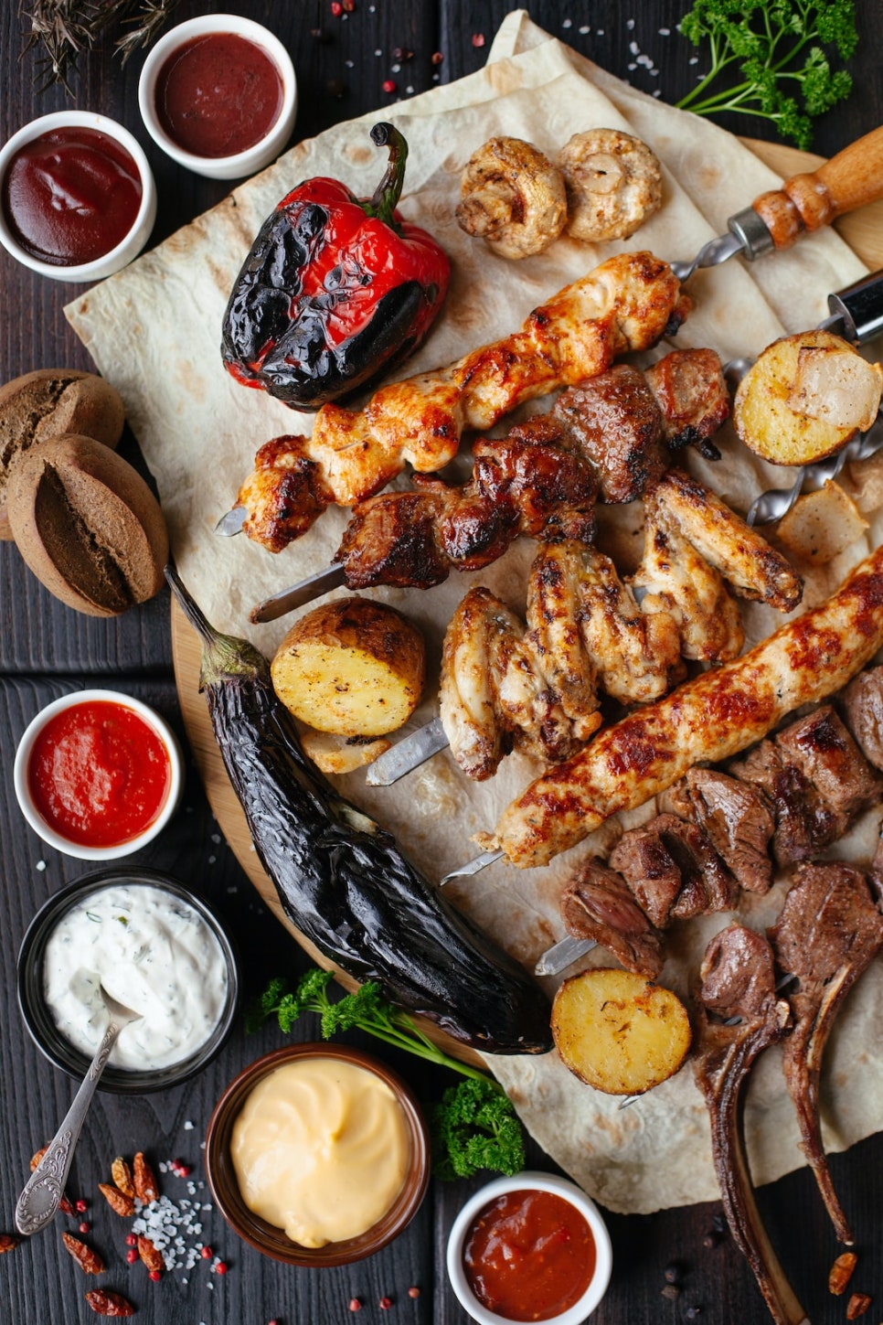 나무 도마 위에 구운 고기 꼬치와 가지, 파프리카, 감자 등 구운 채소와 여러 종류의 소스가 차려져 있습니다.