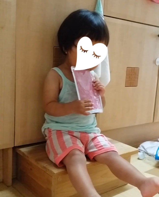 어린아이가 나무 발판에 걸터앉아 양손으로 한약 파우치를 들고 마시고 있는 모습