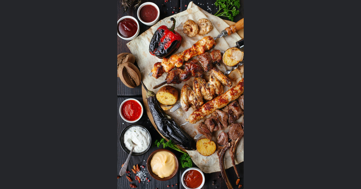 나무 도마 위에 구운 고기 꼬치와 가지, 파프리카, 감자 등 구운 채소와 여러 종류의 소스가 차려져 있습니다.