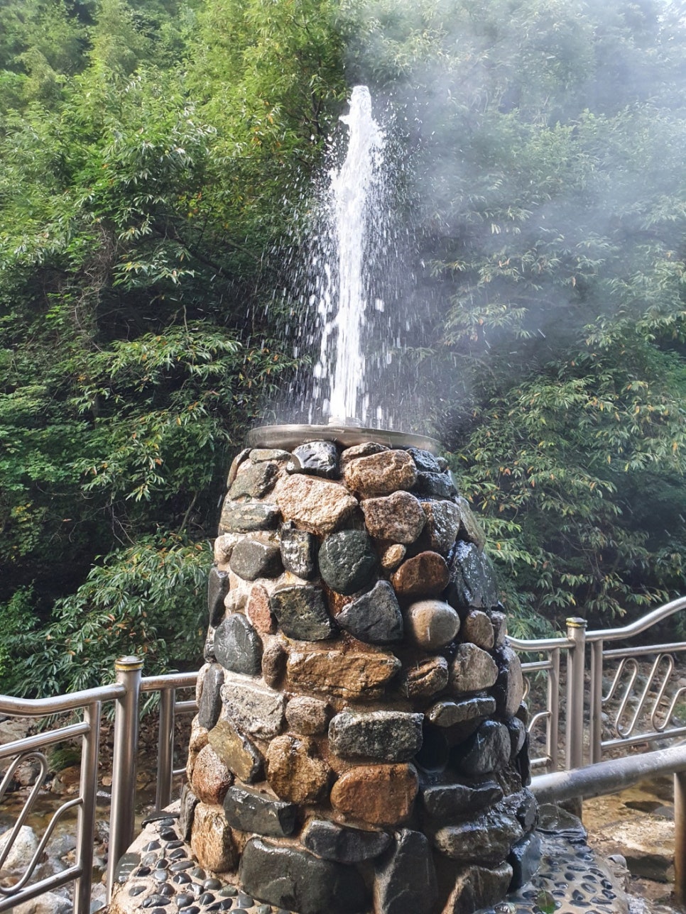 울창한 숲을 배경으로 돌을 쌓아 만든 탑에서 뜨거운 물줄기와 김이 위로 솟구쳐 오르는 모습.